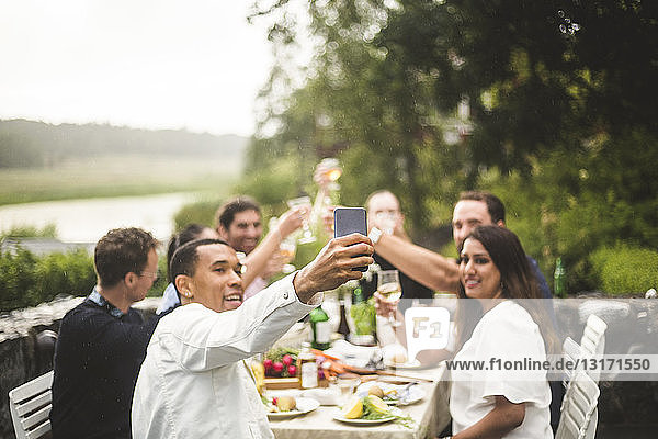 Mann nimmt Selfie mit Freunden und hält Getränke während einer Dinnerparty im Hinterhof