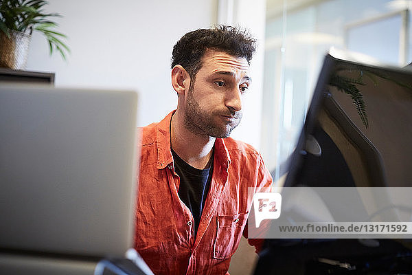 Junger kreativer Geschäftsmann macht ein Gesicht  während er am Computerschreibtisch im Büro arbeitet