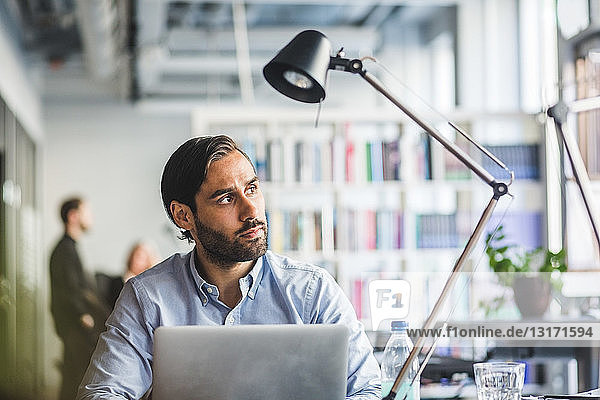 Thoughtful businessman sitting with laptop at desk in creative office