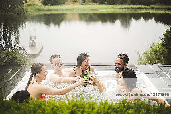 Cheerful male and female friends toasting drinks in hot tub against lake during weekend getaway
