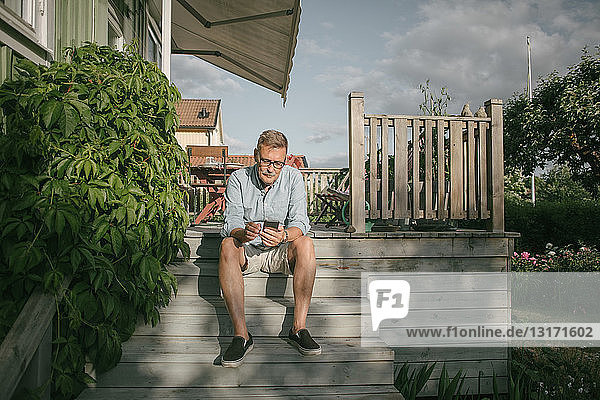 Ein älterer Mann in voller Länge benutzt ein Mobiltelefon  während er auf einer Treppe auf der Veranda sitzt