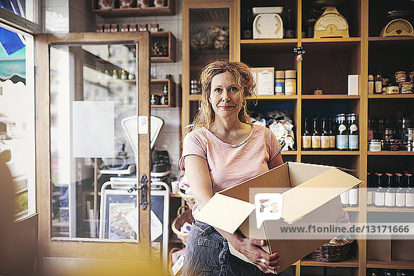 Portrait of confident saleswoman carrying cardboard box against rack in deli