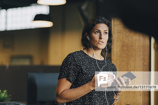 Female executive holding coffee cup while talking with earphones during phone call in office