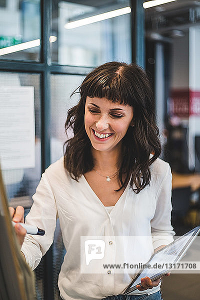 Lächelnde Geschäftsfrau hält digitales Tablett in der Hand  während sie im Büro auf einer Tafel schreibt