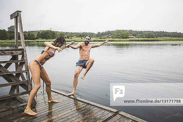 Verspielte Frau in voller Länge schiebt einen männlichen Freund in den See  während sie während eines Wochenendausflugs auf einem Steg steht