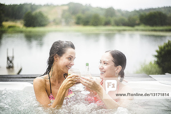 Smiling female friends toasting wineglasses while enjoying in hot tub against lake