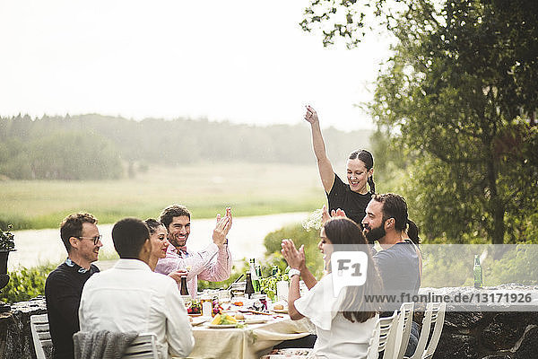 Mid adult woman standing with arm raised while friends applauding at table during dinner party in backyard
