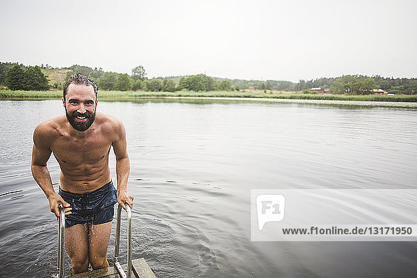 Porträt eines lächelnden Mannes ohne Hemd,  der während eines Wochenendausflugs auf einer Leiter am Steg über dem See steht
