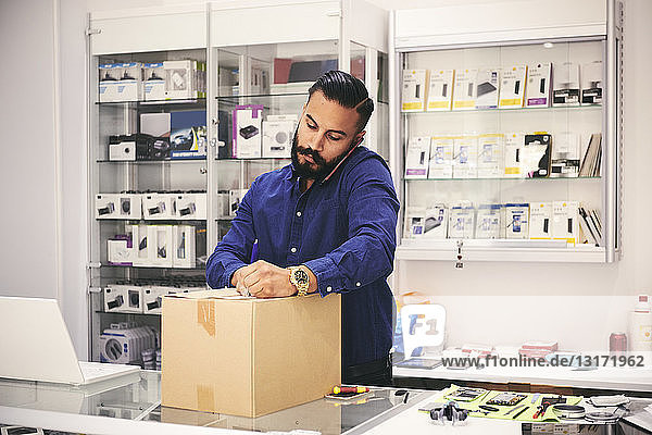 Male owner talking on mobile phone while packing box at counter in electronics store