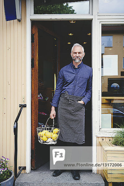 Porträt eines selbstbewussten Verkäufers  der einen Obstkorb hält  während er am Eingang des Geschäfts steht