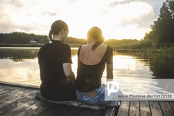 Rear view of female friends sitting on jetty over lake during sunset