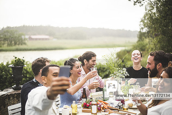Man taking selfie of friends enjoying dinner at table during party in backyard