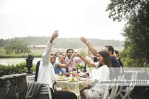 Lächelnder Mann nimmt sich mit Freunden ein Selfie und hält während der Dinnerparty im Hinterhof Getränke in der Hand