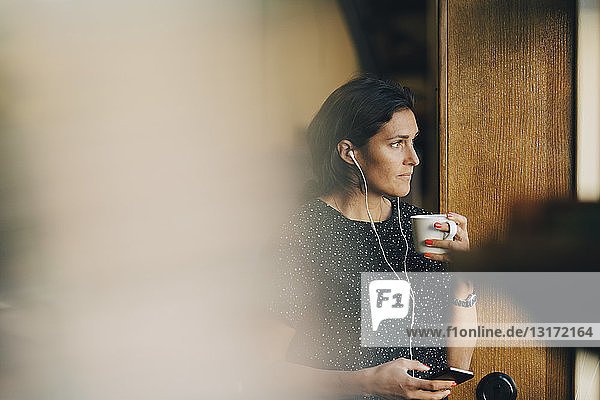 Nachdenkliche Geschäftsfrau  die Kaffee trinkt  während sie im Büro ein Mobiltelefon benutzt