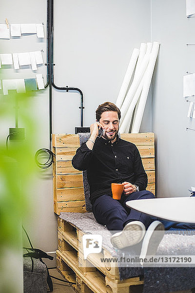 Kreativer Geschäftsmann spricht per Mobiltelefon  während er im Büro auf einem Palettenstapel sitzt