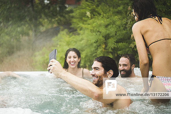 Young man taking selfie through mobile phone while friends enjoying in hot tub during weekend getaway