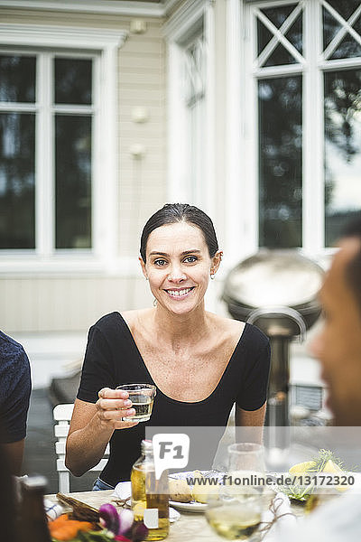 Porträt einer lächelnden Frau mittleren Alters  die ein Getränk im Glas hält  während sie am Esstisch vor der Villa sitzt