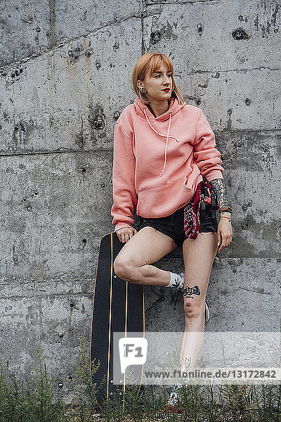 Porträt einer coolen jungen Frau mit Carver-Skateboard  das gegen eine Betonwand gelehnt ist