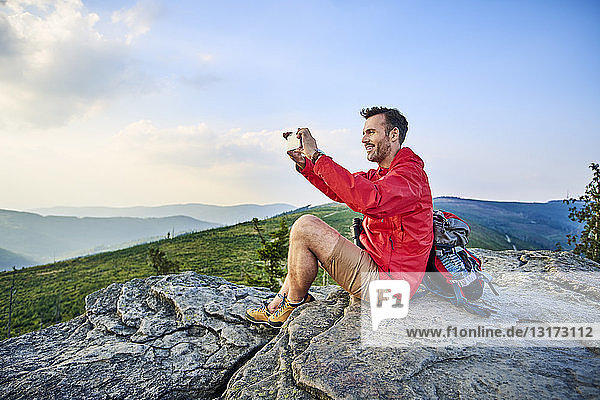 Man sitting on rock taking picture with his cell phone during hiking trip in the mountains