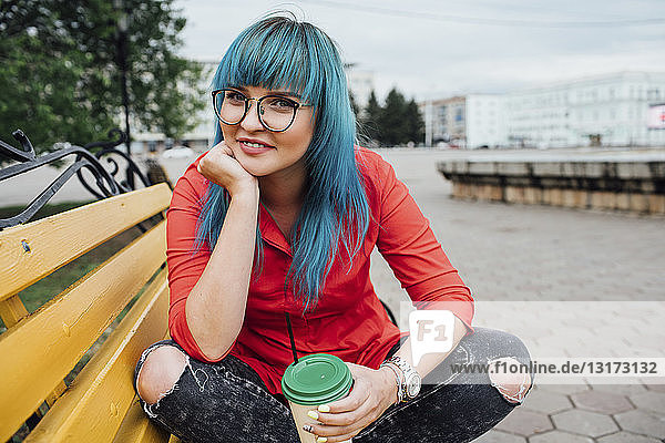 Porträt einer lächelnden jungen Frau mit blau gefärbten Haaren  die mit einem Getränk auf einer Bank sitzt
