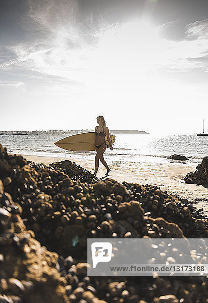Frankreich  Bretagne  junge Frau trägt Surfbrett an einem felsigen Strand am Meer