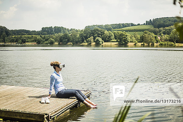 Frau mit VR-Brille sitzt auf einem Steg an einem See mit Füßen im Wasser
