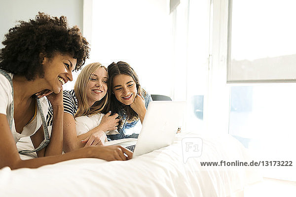 Three happy girlfriends lying on bed sharing laptop