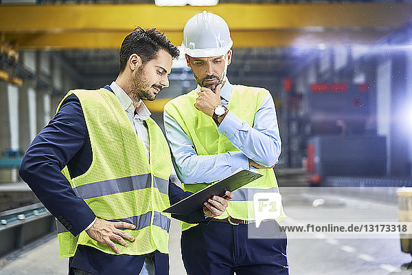 Zwei Manager  die Arbeitsschutzkleidung tragen  schauen in der Fabrik auf die Zwischenablage