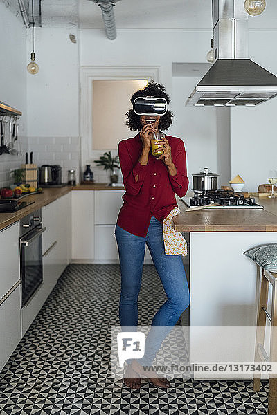 Frau steht in ihrer Küche  trägt VR-Brille  trinkt Orangensaft