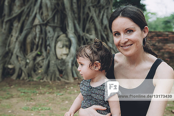 Thailand  Ayutthaya  Porträt einer lächelnden Mutter und Tochter  die den Buddhakopf zwischen Baumwurzeln im Wat Mahathat besuchen