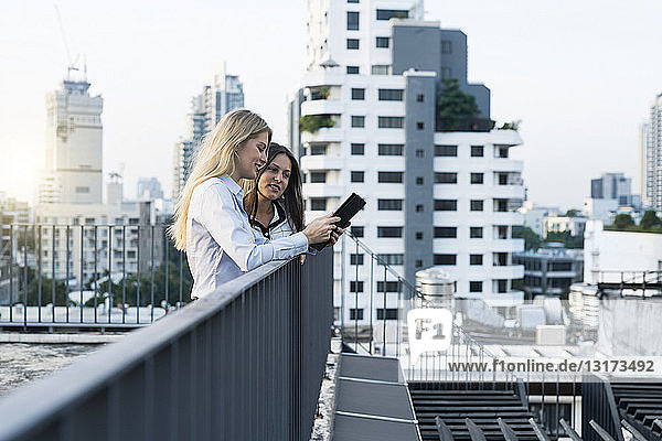 Two businesswomen talking on city rooftop  using digital tablet