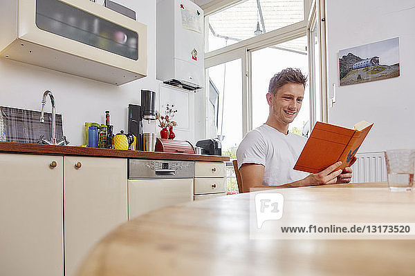 Lächelnder junger Mann sitzt zu Hause am Tisch und liest ein Buch