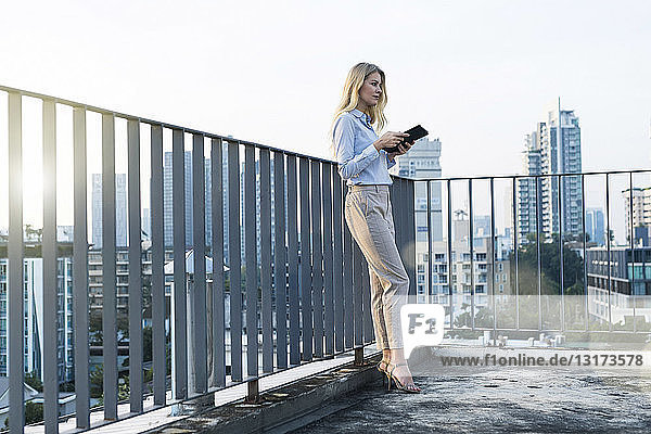 Blonde business woman leaning onto handrail holding tablet on city rooftop