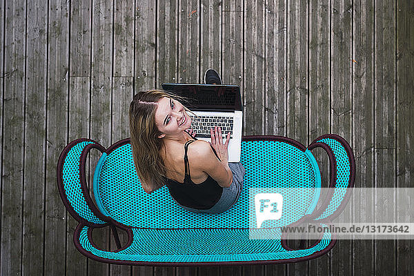 Lächelnde junge Frau sitzt auf türkisfarbener Couch auf der Terrasse mit Laptop  Draufsicht