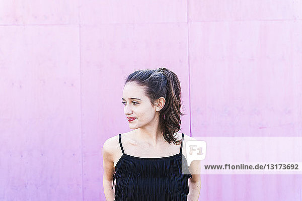 Lächelnde junge Frau vor rosa Wand mit seitlichem Blick