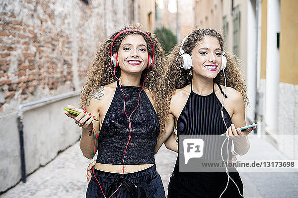 Portrait of smiling twin sisters listening music with headphones and cell phones in the city