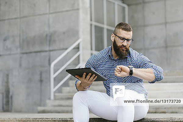 Porträt eines bärtigen Hipster-Geschäftsmannes auf einer Treppe sitzend mit digitalem Tablett  das die Zeit überprüft