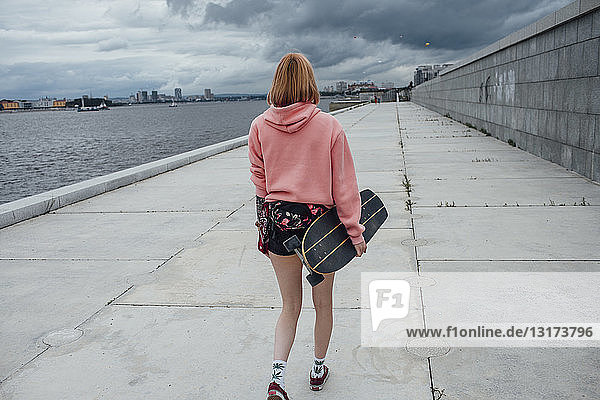 Rückansicht einer jungen Frau  die ein Carver-Skateboard hält und am Flussufer spazieren geht