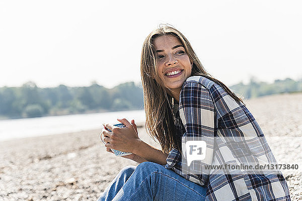 Glückliche junge Frau mit einem Becher in der Hand am Flussufer sitzend