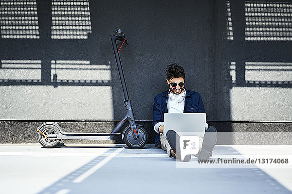 Entspannter junger Mann sitzt neben seinem Elektroroller auf dem Boden und benutzt seinen Laptop