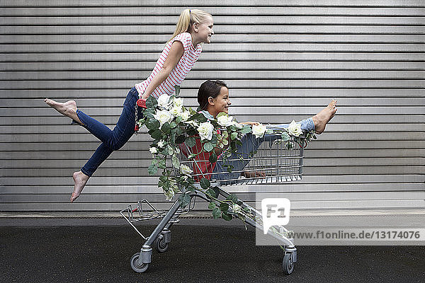 Zwei Mädchen spielen mit einem Einkaufswagen  der mit weißen Kunstblumen geschmückt ist