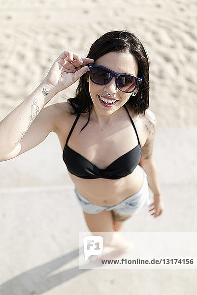 Porträt einer lächelnden jungen Frau mit Nasenpiercing und Tätowierungen mit Sonnenbrille am Strand