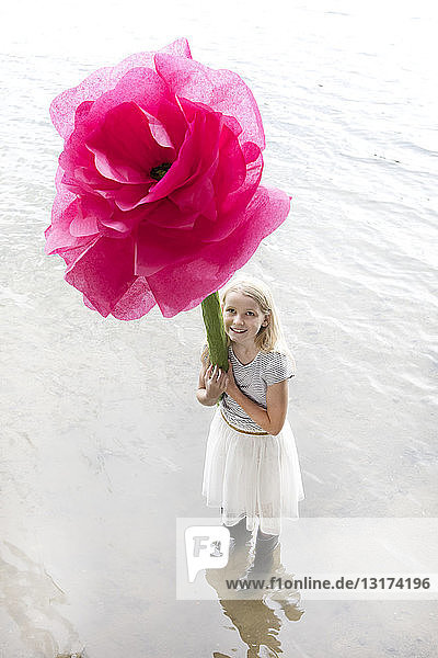 Porträt eines lächelnden blonden Mädchens  das in einem See steht und eine überdimensionale rosa Kunstblume hält