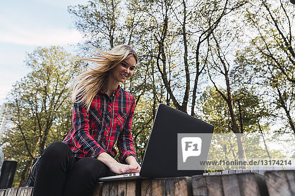 Porträt einer lächelnden jungen Frau  die auf einer Bank im Freien sitzt und am Laptop arbeitet