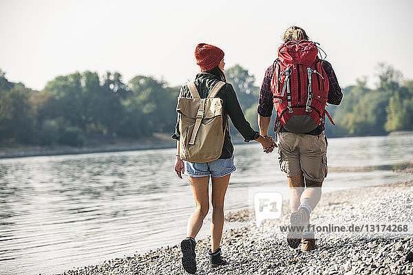 Rückansicht eines jungen Paares mit Rucksäcken  das Hand in Hand am Flussufer geht