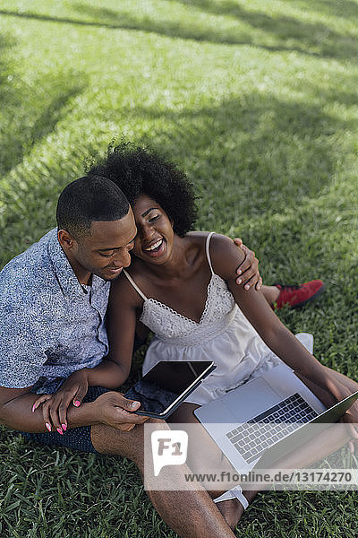 Glückliches junges Paar mit Tablett und Laptop auf einem Rasen in einem Park