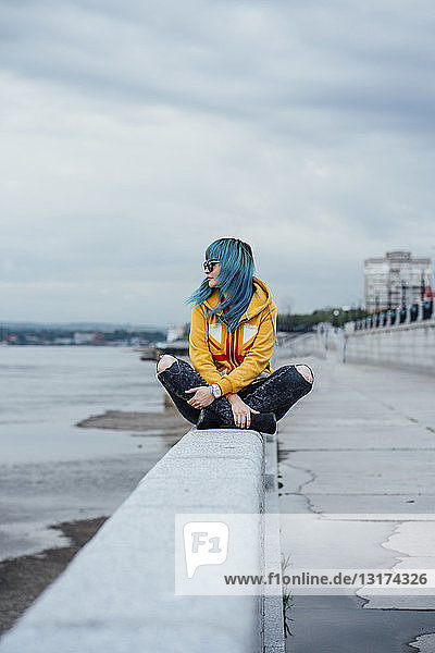 Young woman with dyed blue hair sitting on a wall looking at distance