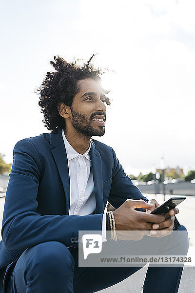 Lächelnder junger Geschäftsmann sitzt draußen und hält sein Handy