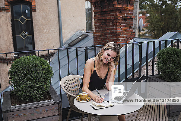 Lächelnde junge Frau sitzt mit Laptop auf dem Balkon und macht sich Notizen