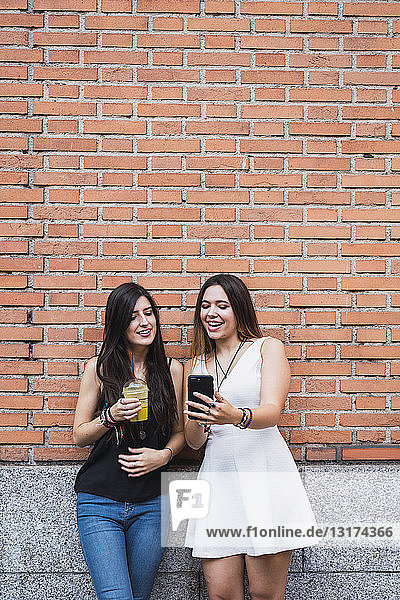 Mädchen-Freunde stehen vor einer Backsteinmauer und schauen auf ein Smartphone